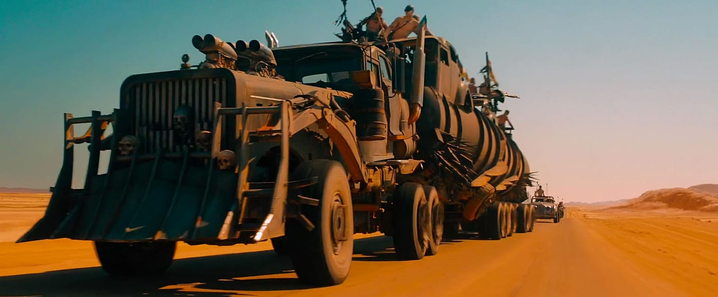 The War Rig from Mad Max: Fury Road rolls out across orange desert, escort cars trailing behind.