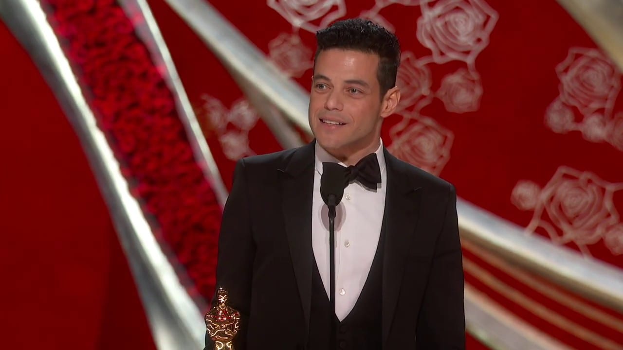Rami Malek at the Oscars podium in a tuxedo, smiling mid-speech with his Best Actor statuette.