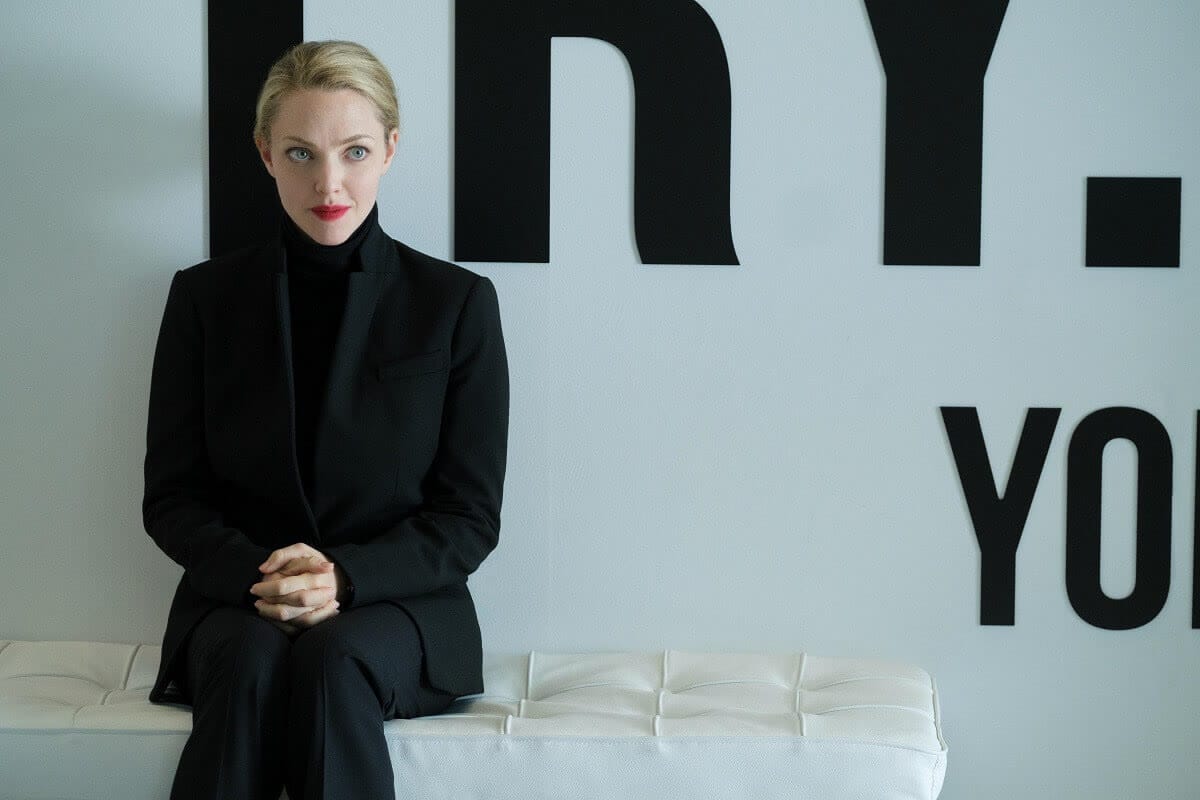 Amanda Seyfried as Elizabeth Holmes in The Dropout, in a black turtleneck, sitting on a white bench.